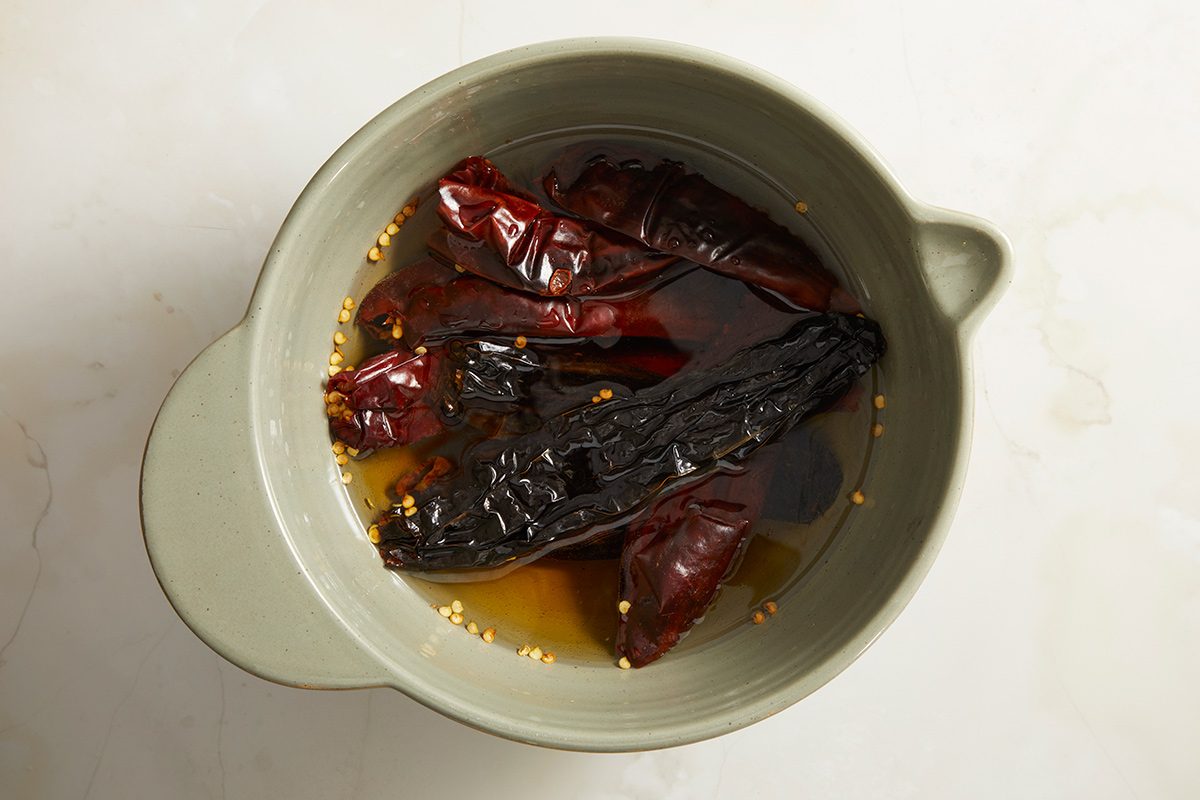 A light green bowl containing dried red and dark brown chiles soaking in water, with some seeds floating around them, on a light-colored surface.