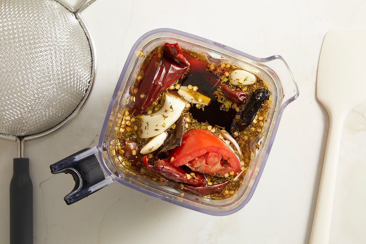 A blender container filled with dried red chilies, garlic, tomato, seeds, and dark liquid sits on a white surface next to a metal strainer and a white spatula.