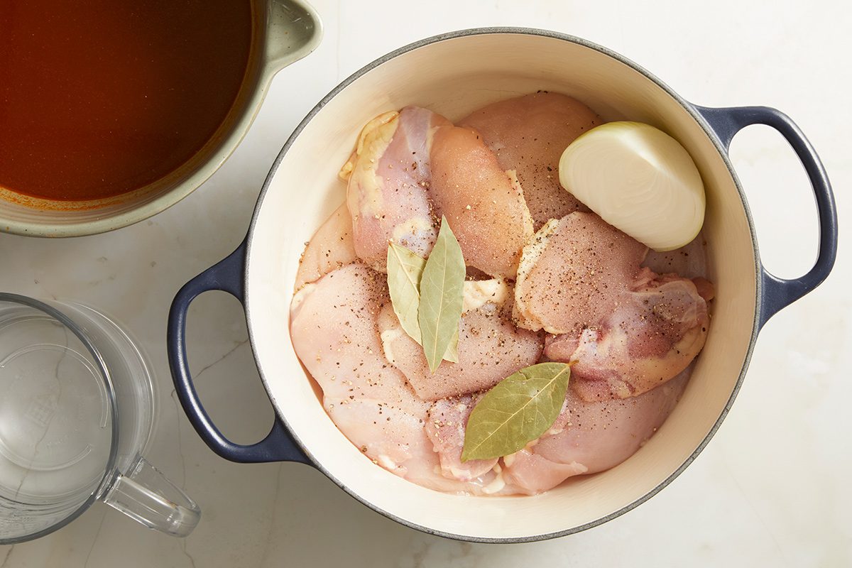 Raw chicken pieces, bay leaves, and a halved onion in a blue Dutch oven pot, ready for cooking. A glass measuring cup and a bowl of reddish broth are beside the pot on a white surface.