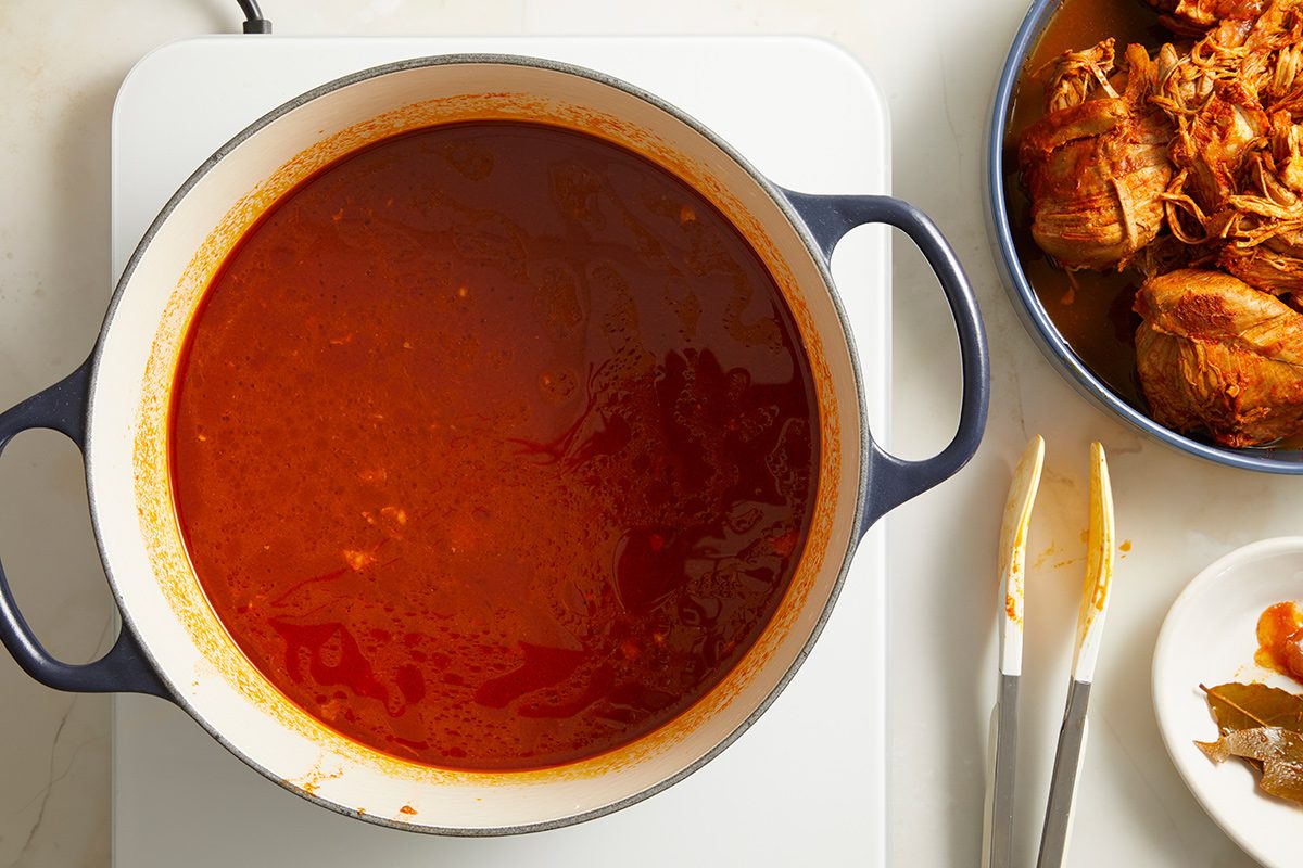 A pot of rich red sauce sits on a stovetop next to a bowl of shredded meat, metal tongs, and a fork on a white surface.