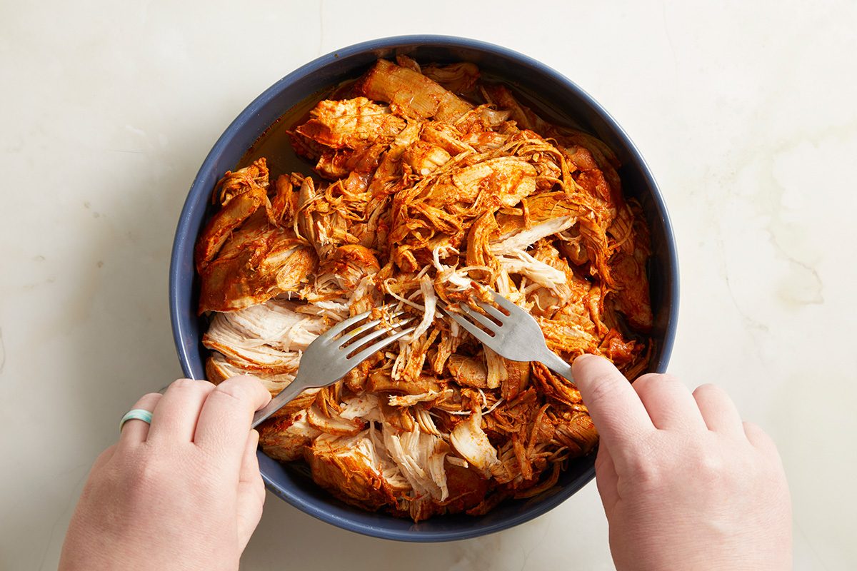 Two hands use forks to shred cooked, seasoned chicken in a dark bowl on a light surface.