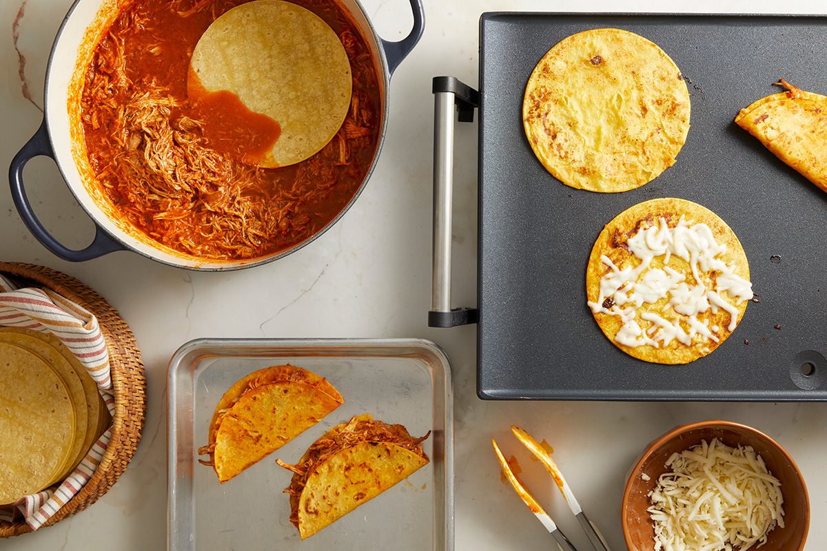 Overhead view of assembling tacos: a pot of shredded meat in red sauce, tortillas being dipped and cooked on a griddle, one taco filled with meat and cheese, and bowls of tortillas and shredded cheese nearby.