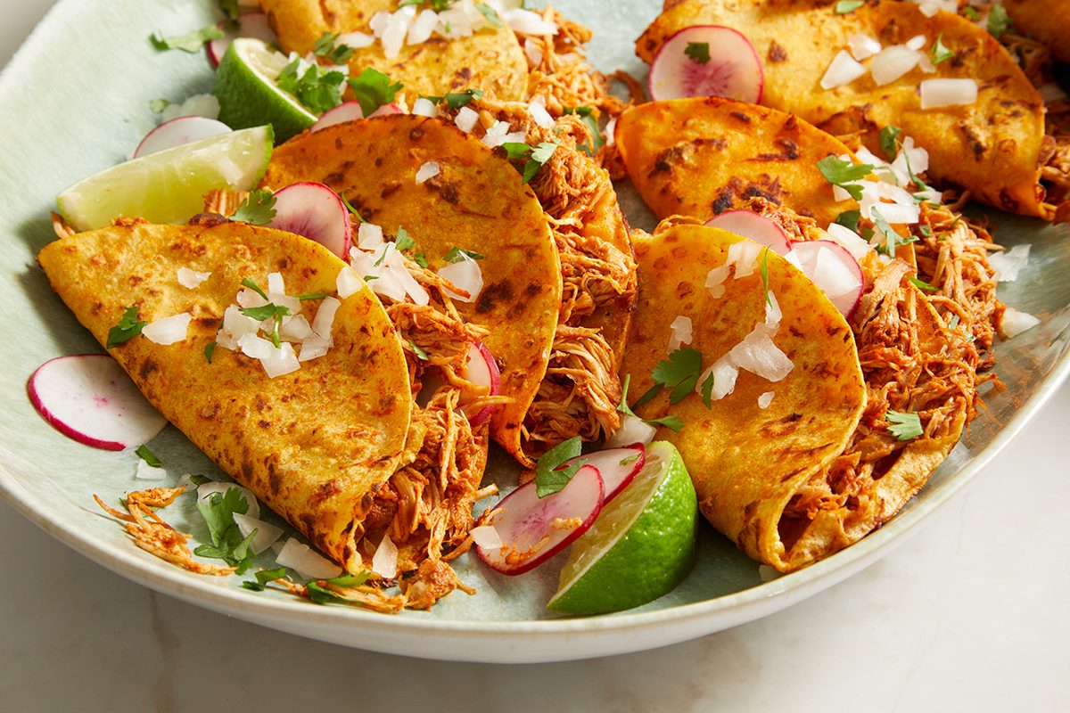 Three crispy shredded chicken tacos topped with chopped onions, cilantro, and sliced radishes are arranged on a plate with lime wedges.