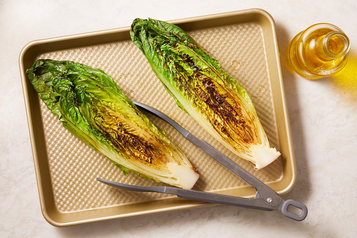 Overhead horizontal image of halved romaine lettuce being grilled or charred on a sheet pan with olive oil.