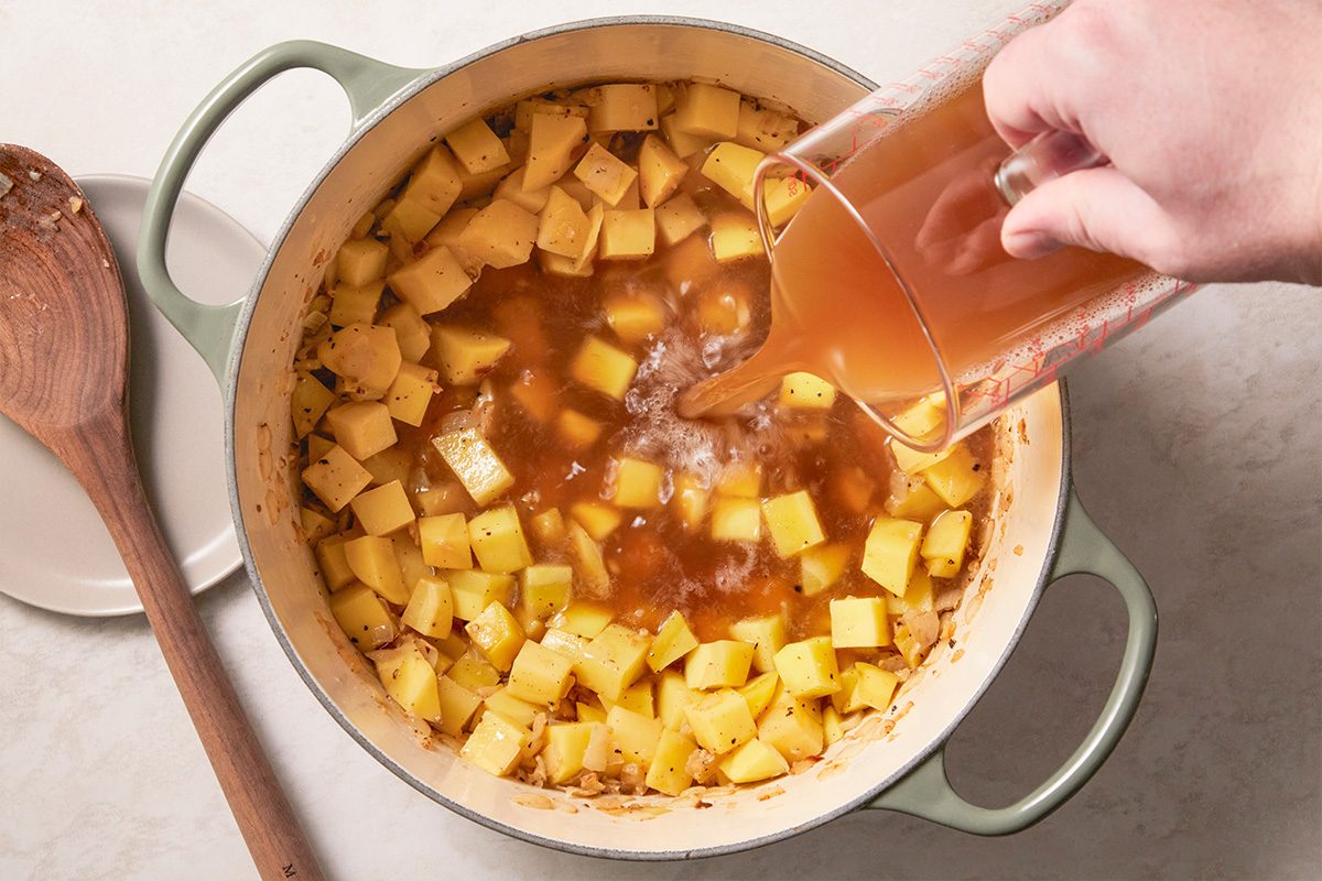 Overhead horizontal image of diced potatoes simmering in broth as liquid is being poured into a Dutch oven.