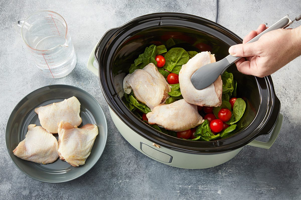 Overhead horizontal image of raw chicken thighs placed into a slow cooker over fresh spinach and cherry tomatoes.