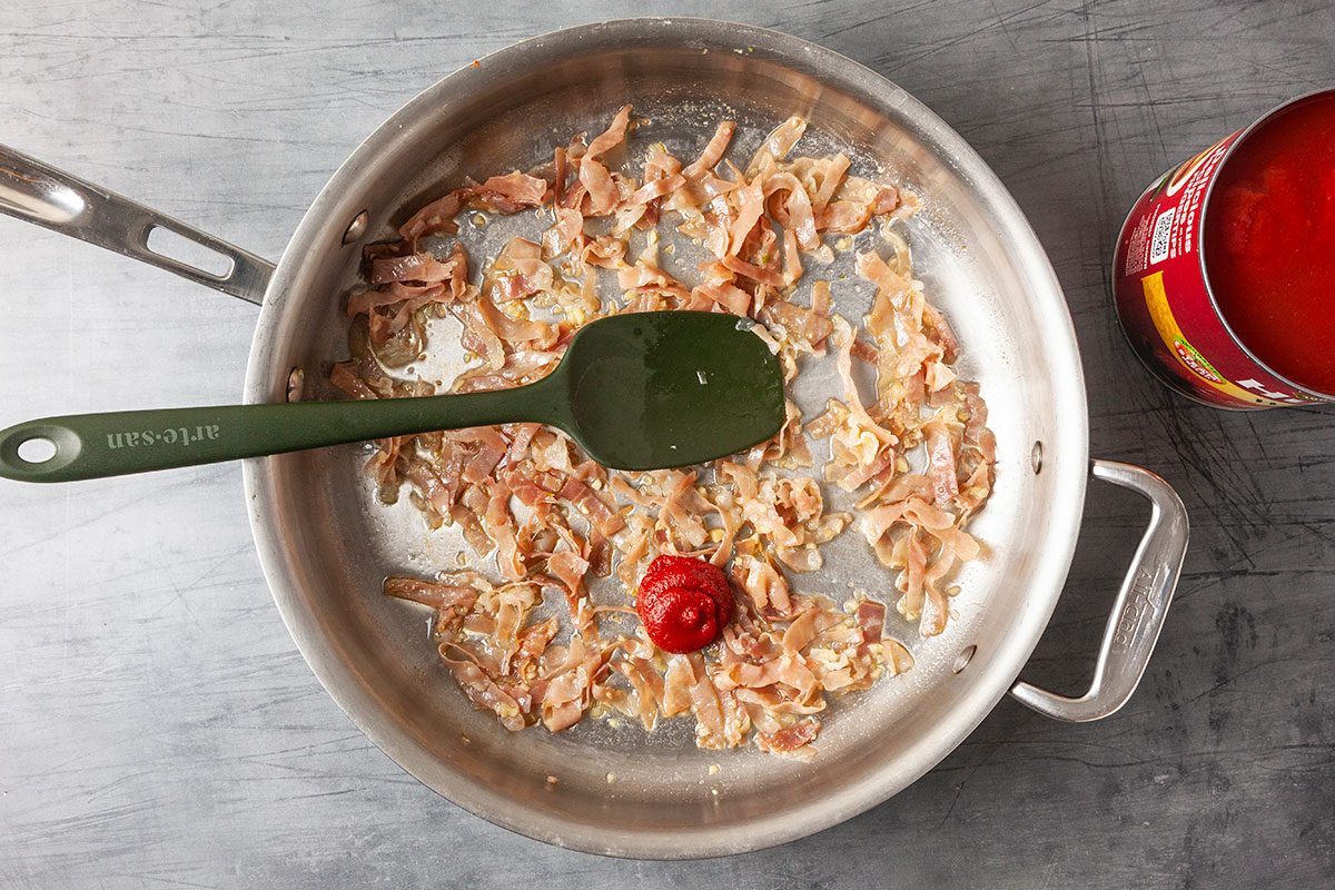 Overhead view of butter melting in a skillet with minced garlic and thinly sliced prosciutto beginning to sauté, marking the start of a homemade vodka sauce for Chicken Parmesan. Ingredients are gently cooking in a light-colored pan on a gray surface in this step-by-step preparation image.