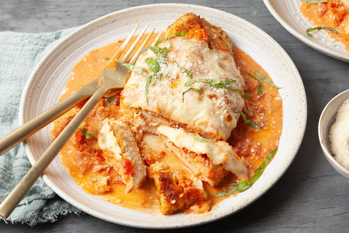 Close-up serving shot of Chicken Parmesan with vodka sauce as a fork cuts into the crispy breaded chicken, revealing tender interior beneath melted mozzarella and creamy sauce.