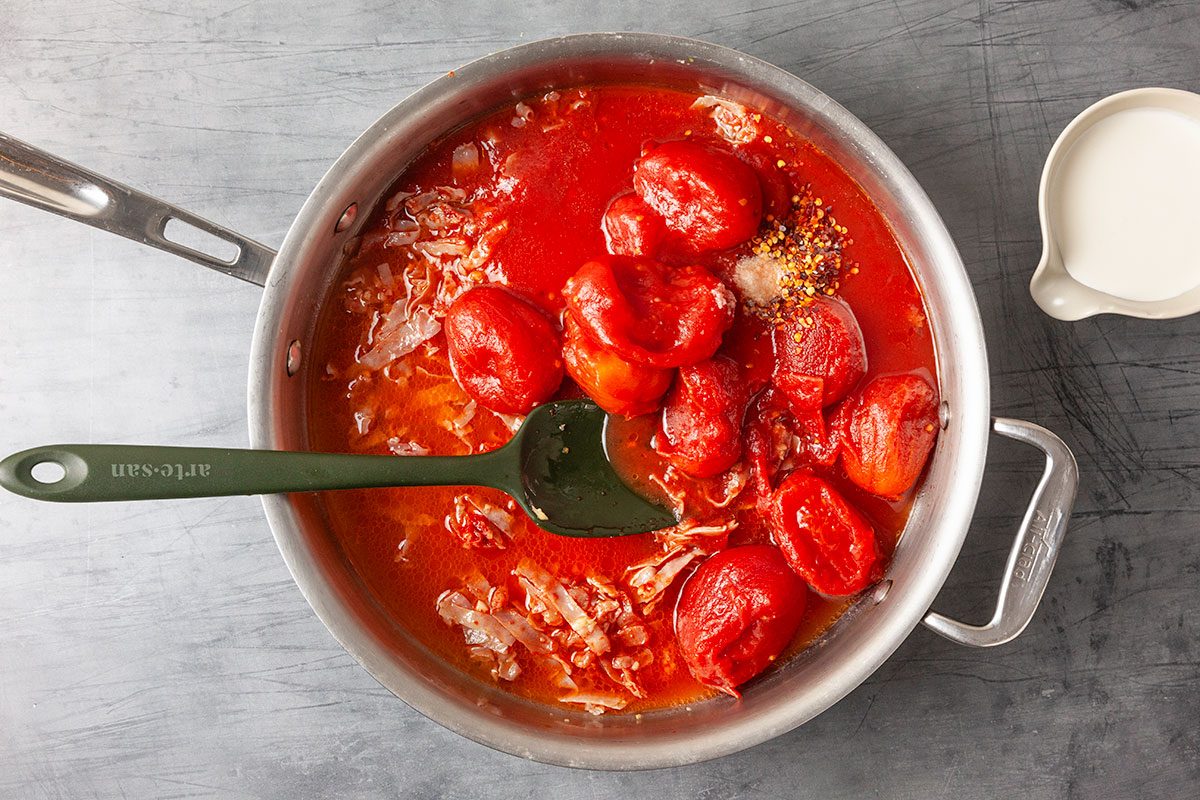 Overhead process image of whole plum tomatoes added to sautéed garlic and prosciutto in a skillet as the vodka sauce develops. A green spatula rests in the pan while the tomatoes simmer in butter.