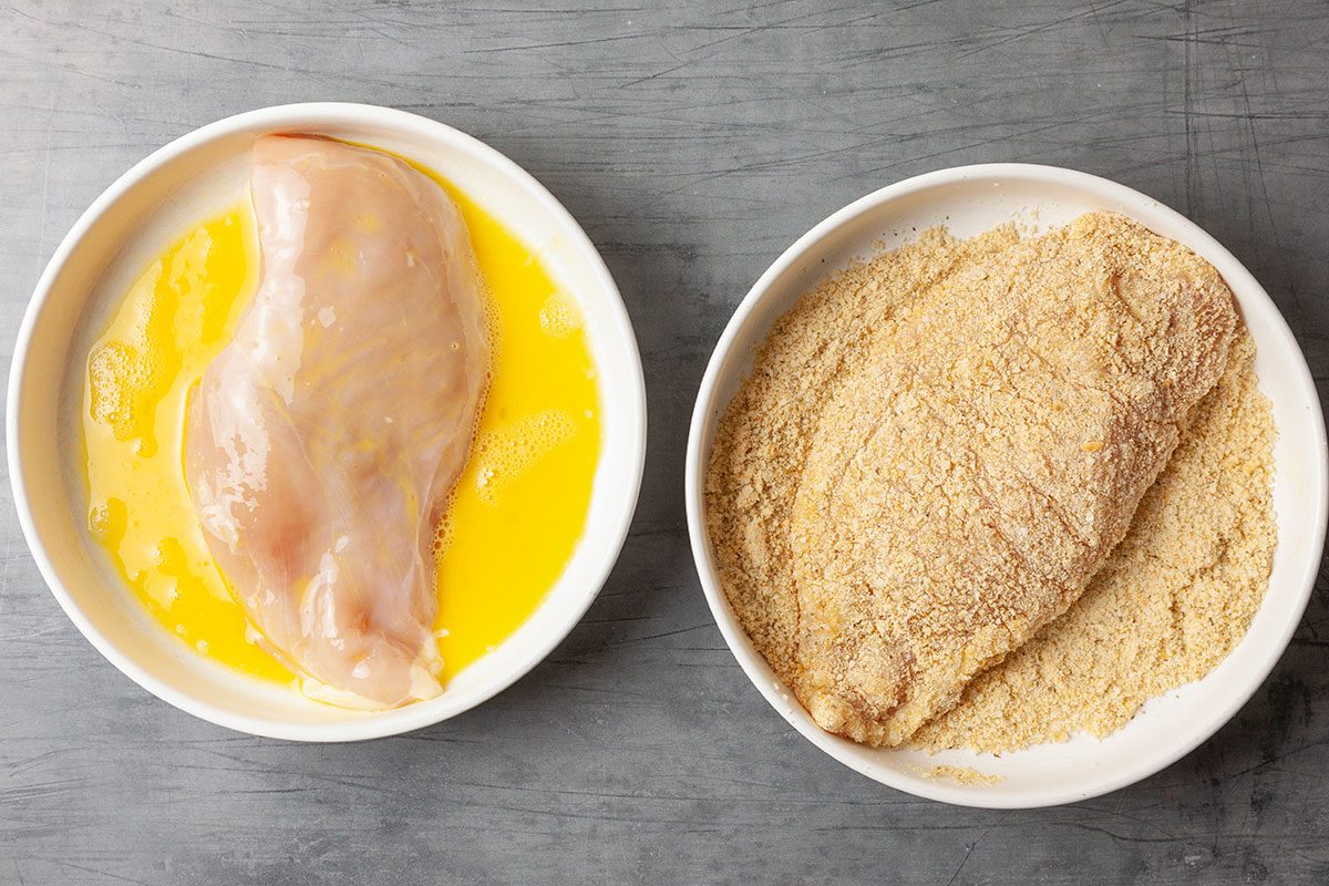 Overhead preparation shot of breading station with raw chicken breast dipped in beaten egg and coated in seasoned bread crumbs and grated Parmesan cheese, ready for pan frying.