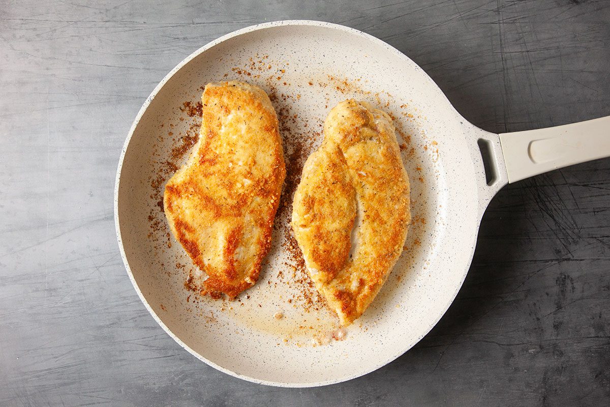 Overhead cooking image of breaded chicken breast halves browning in olive oil in a skillet. The golden crust forms as the chicken cooks evenly on both sides.