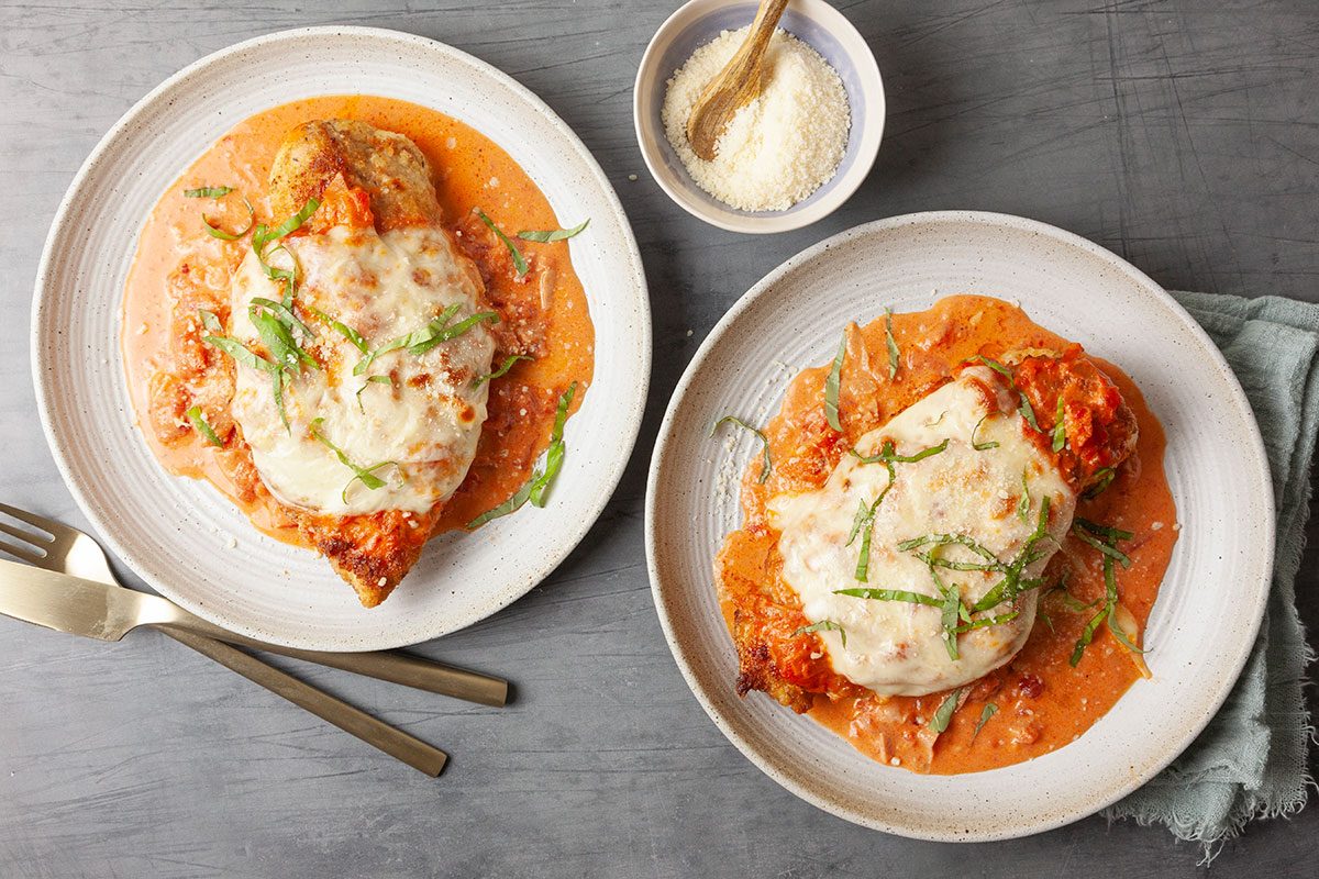 Overhead hero shot of finished Chicken Parmesan with creamy vodka sauce and melted mozzarella, garnished with fresh basil and additional Parmesan cheese, served on white plates.