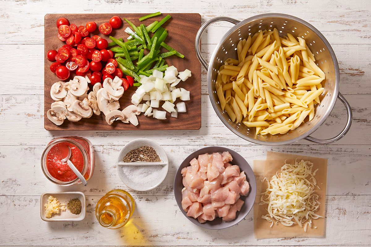 Overhead shot of ingredients for a pasta dish on a white wooden surface, including cooked penne, chopped vegetables, diced chicken, shredded cheese, seasonings, olive oil, minced garlic, and tomato sauce;