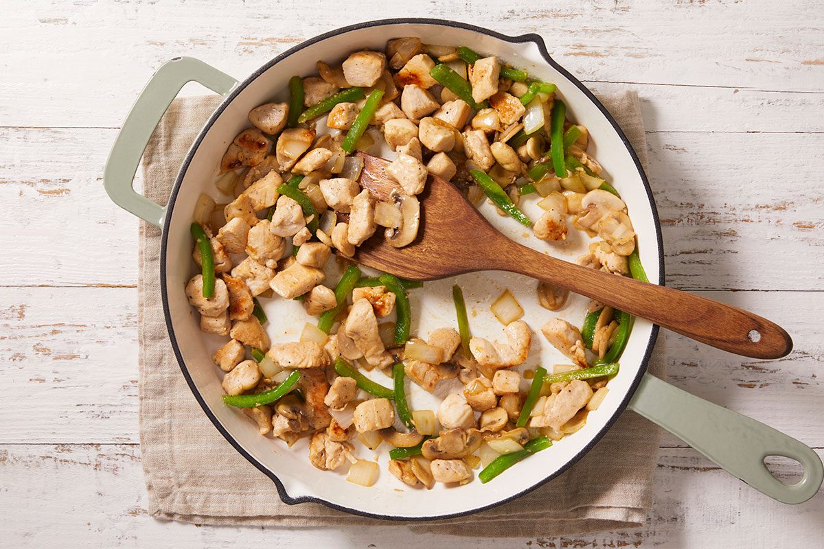 Overhead shot of a skillet with cooked chicken pieces, sliced green bell peppers, and onions being stirred with a wooden spoon on a tan cloth over a white wooden surface;