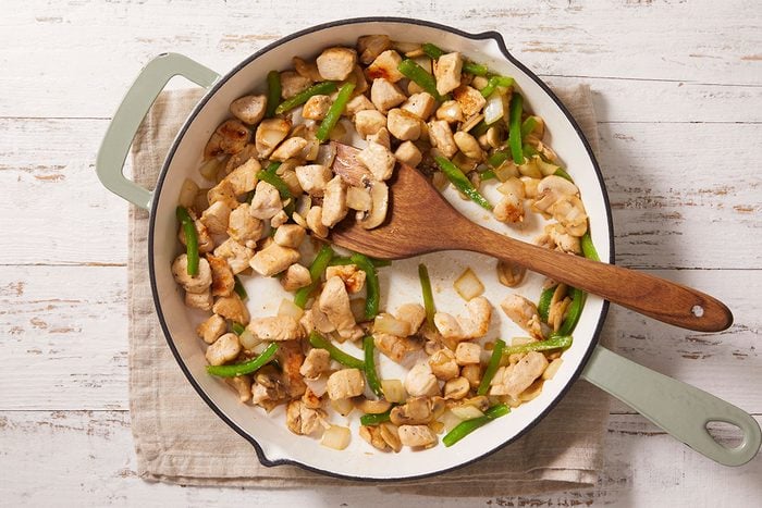 Overhead shot of a skillet with cooked chicken pieces, sliced green bell peppers, and onions being stirred with a wooden spoon on a tan cloth over a white wooden surface;