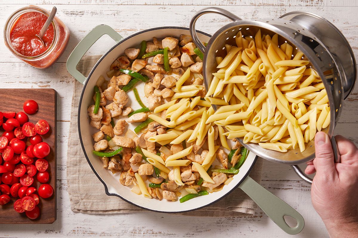 Overhead shot of a person pouring cooked penne from a colander into a pan with sautéed chicken and green bell peppers, with a bowl of tomato sauce and a cutting board of sliced cherry tomatoes nearby;