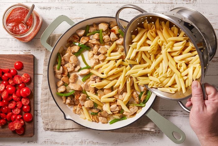 Overhead shot of a person pouring cooked penne from a colander into a pan with sautéed chicken and green bell peppers, with a bowl of tomato sauce and a cutting board of sliced cherry tomatoes nearby;