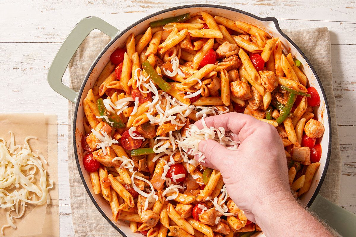 Overhead shot of a hand sprinkling shredded cheese over a pan of cooked penne pasta mixed with chicken, cherry tomatoes, and green bell peppers on a wooden table;