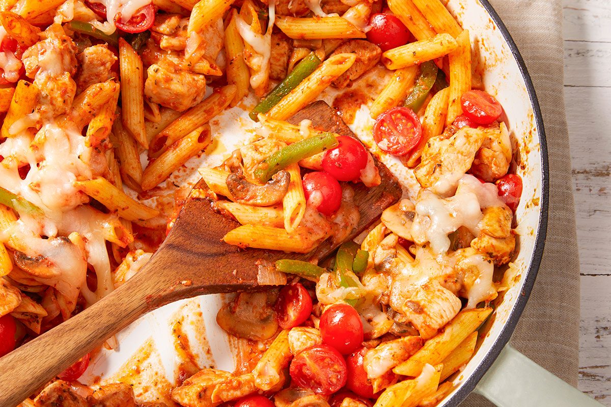 Closeup shot of Italian chicken and penne being stirred with a wooden spoon, with cherry tomatoes, bell peppers, and melted cheese visible;