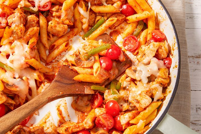 Closeup shot of Italian chicken and penne being stirred with a wooden spoon, with cherry tomatoes, bell peppers, and melted cheese visible;