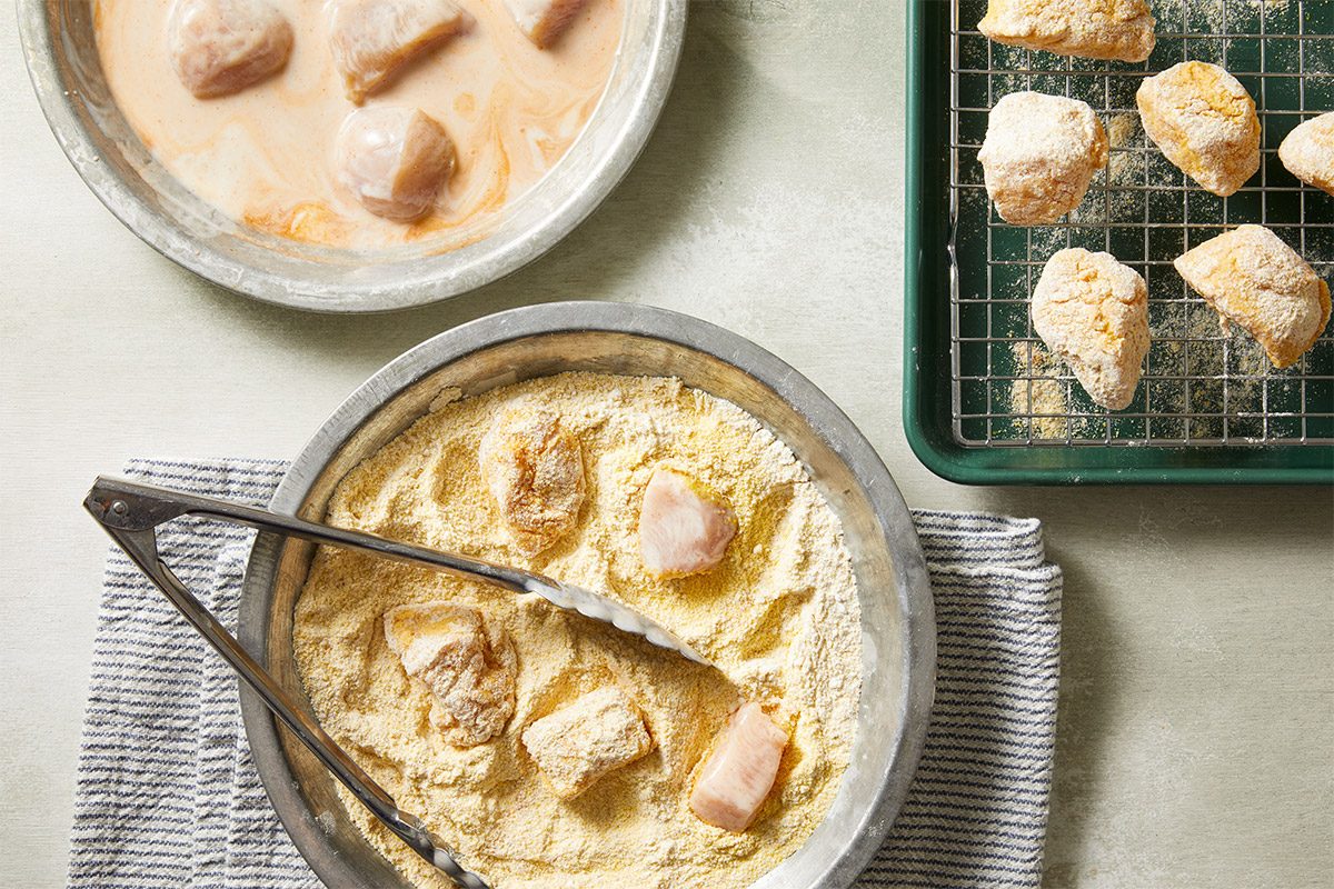 Overhead shot of fried chicken pieces coated in batter resting on a cooling rack beside a bowl of creamy sauce.