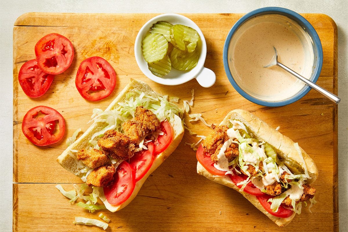 Overhead shot of sandwich ingredients arranged on a cutting board including tomato slices, pickles, shredded lettuce, and a sandwich roll.