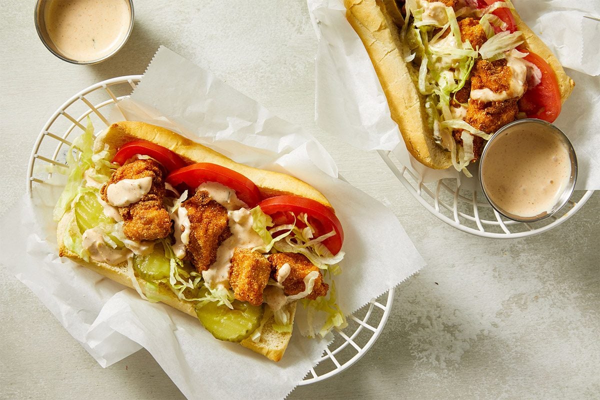 Overhead shot of Chicken Po'Boys sandwiches wrapped in parchment paper, topped with sauce and served with pickles.