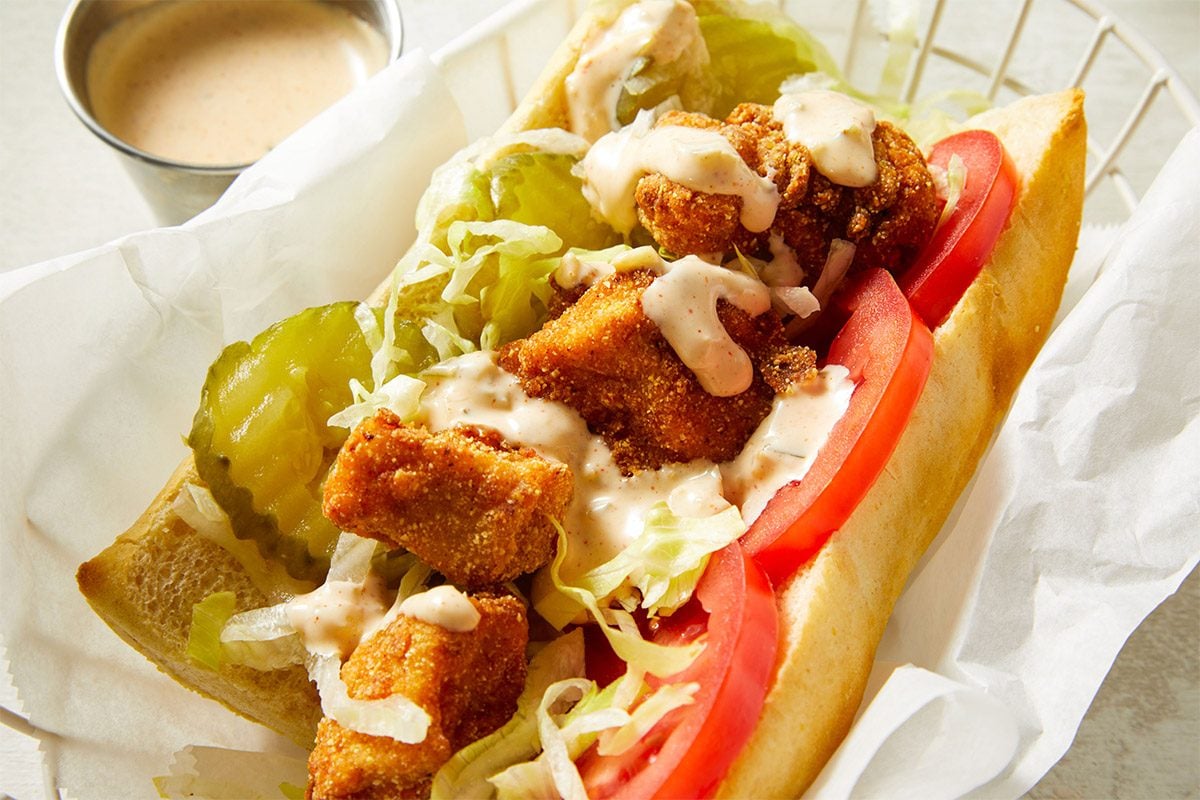 Close-up overhead shot of Chicken Po'Boys sandwiches wrapped in parchment paper, topped with sauce and served with pickles.