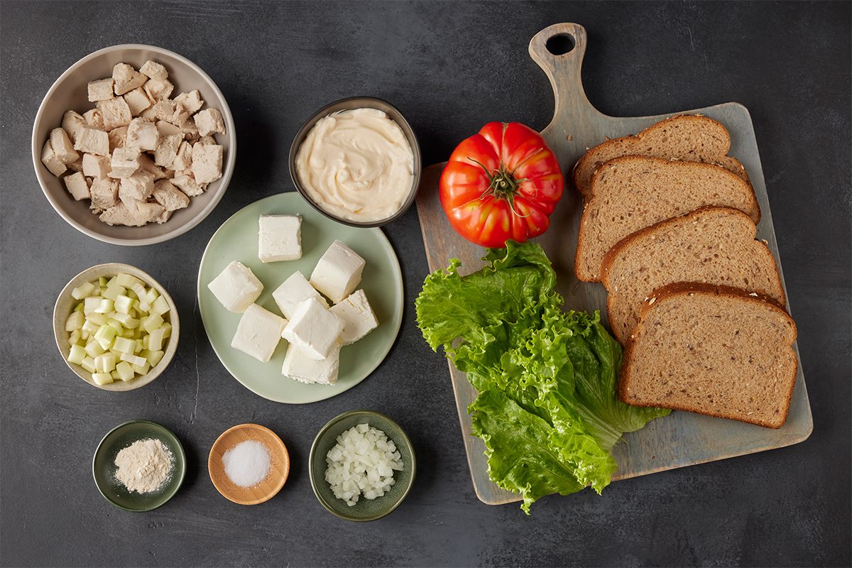 Top view shot of sandwich ingredients—diced chicken, mayonnaise, cubed cheese, chopped celery, chopped onion, lettuce leaves, a tomato, seasonings, and four slices of whole-grain bread arranged on a cutting board; all set on a dark surface;