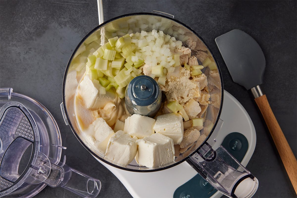 Overhead shot of a food processor bowl filled with chopped celery, onions, cream cheese, mayonnaise, and chunks of cooked chicken, ready to be blended; A spatula rests nearby on the countertop;