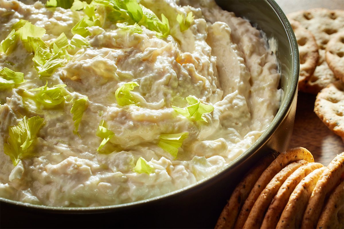 Closeup shot of creamy chicken spread topped with chopped celery in a bowl on a wooden board, surrounded by round crackers;