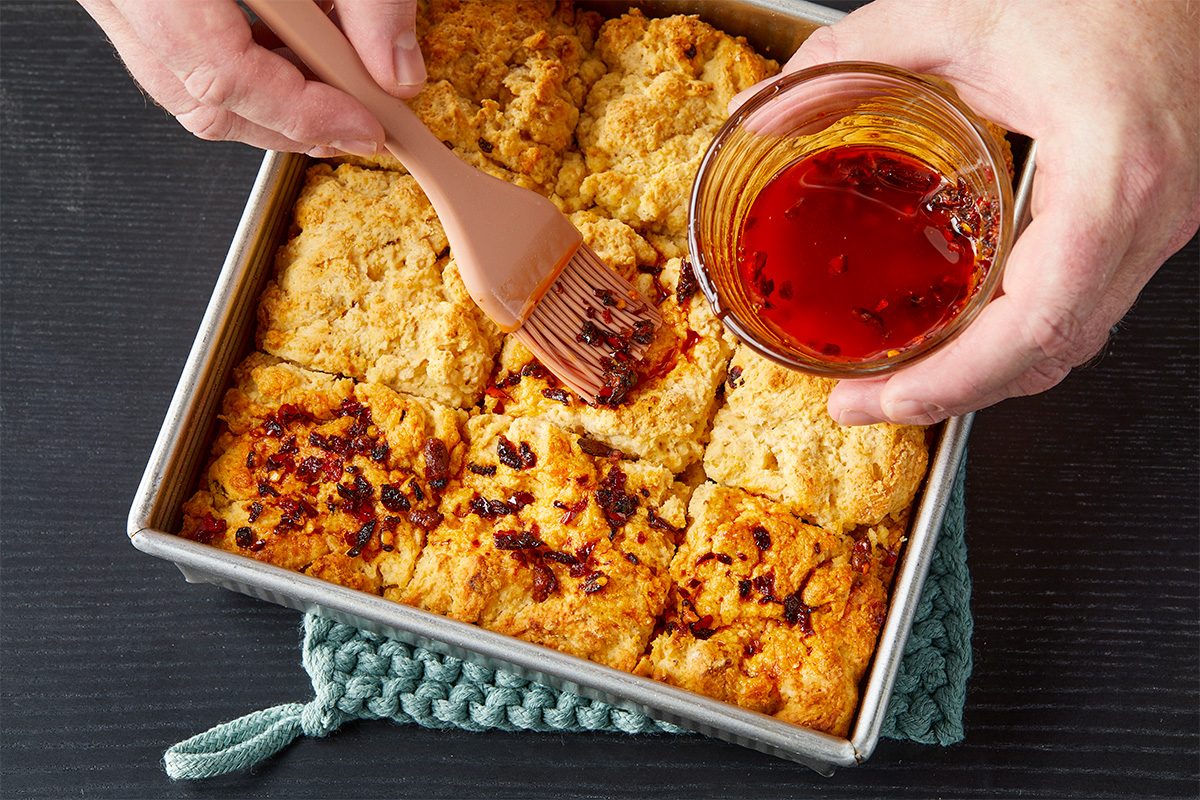 Overhead image of baked chili crisp biscuits in a square baking pan as chili crisp butter is brushed across the golden tops, adding flavor and a glossy finish.
