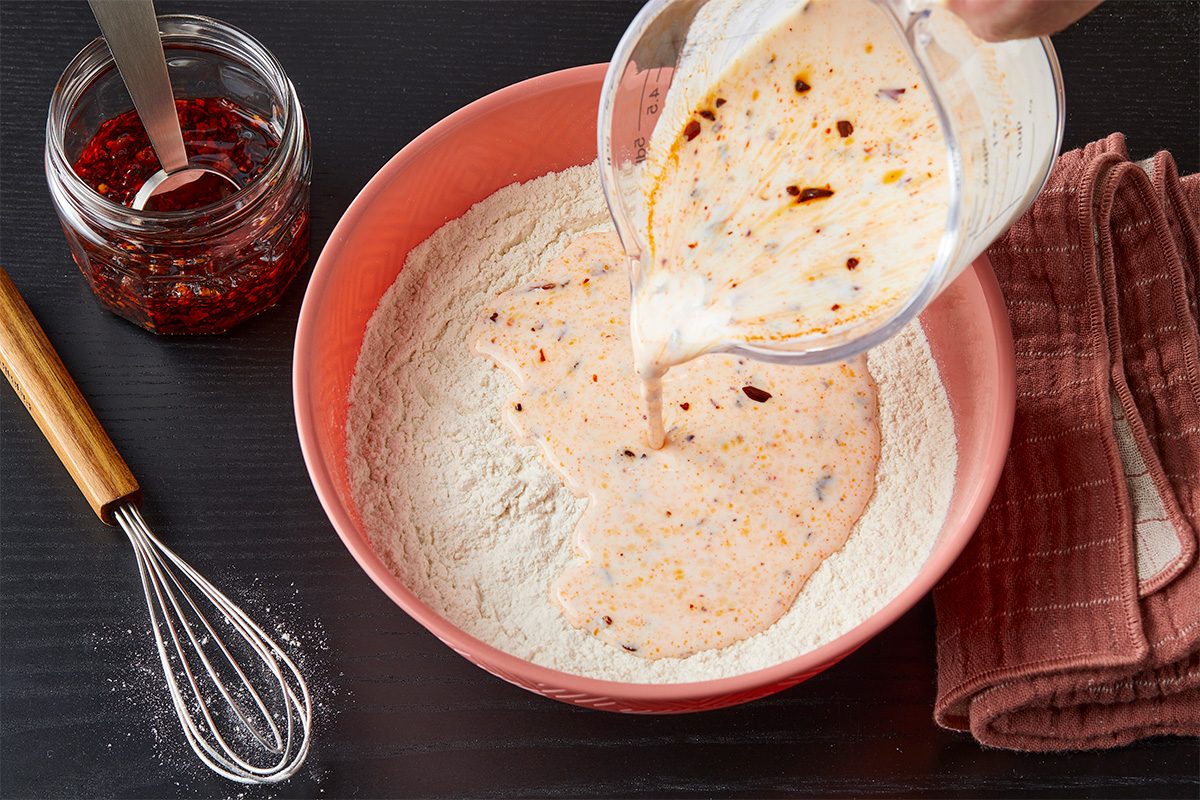 Overhead view of chili crisp buttermilk mixture being poured into a bowl of flour and dry ingredients while preparing biscuit dough.