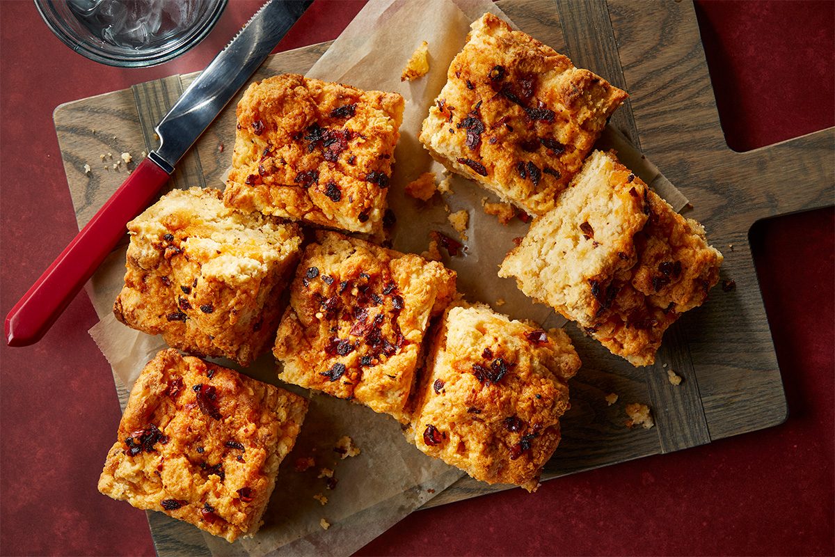 Overhead composition of chili crisp biscuit squares served on a cutting board with a knife, highlighting the golden tops and soft interiors.