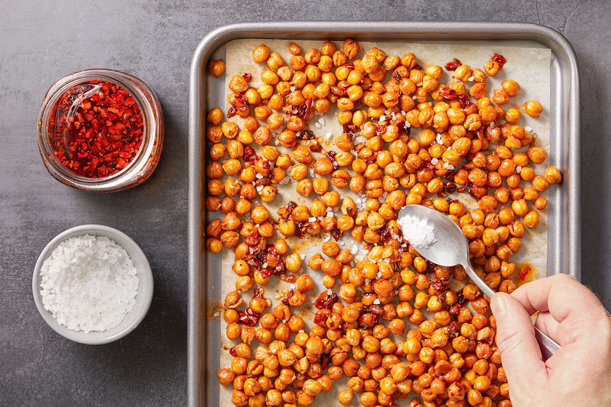 Overhead horizontal image of chickpeas on a sheet pan being seasoned with salt and chili flakes.