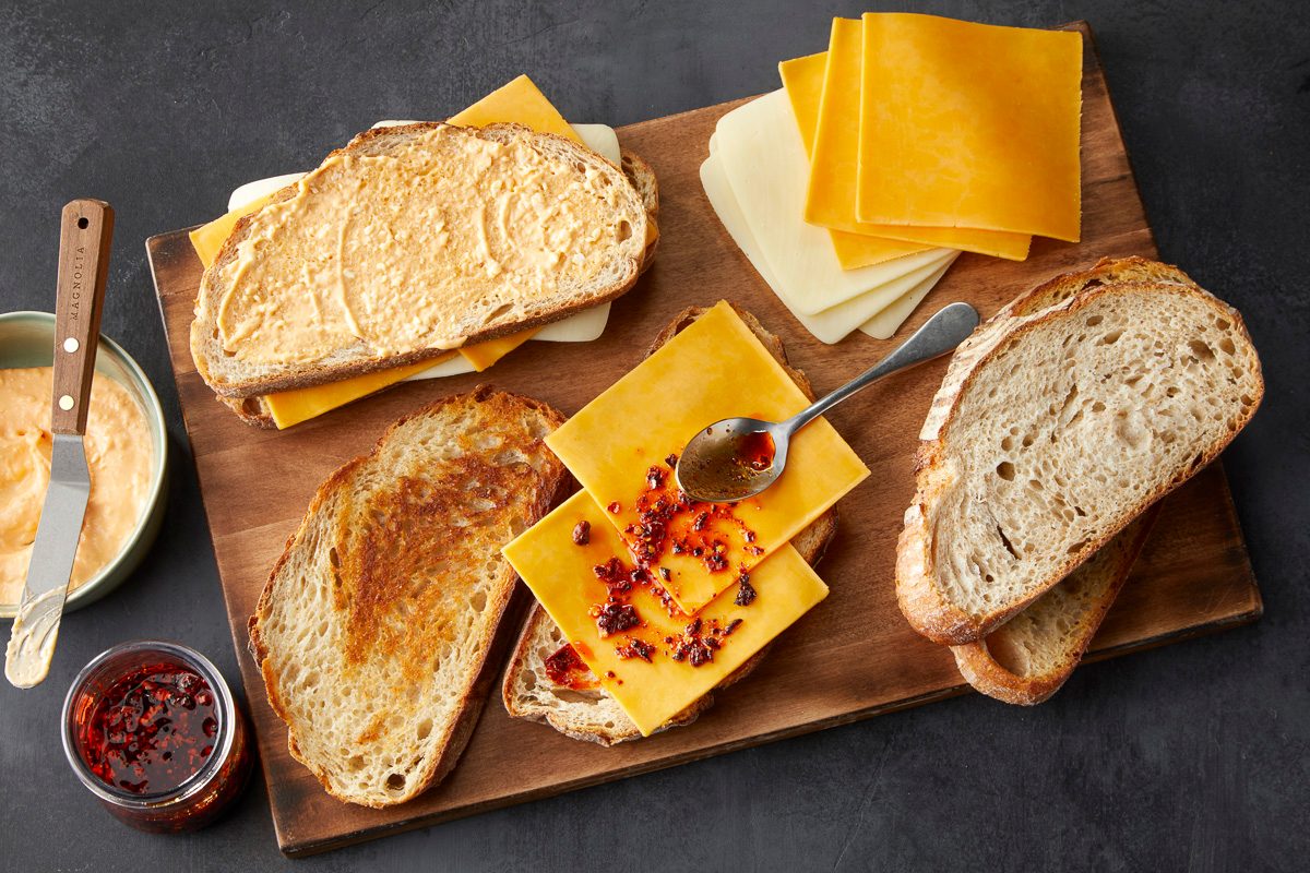 toasted bread topped with cheddar cheese slices drizzled with chili crisp next to bread and slices of cheese on a chopping board