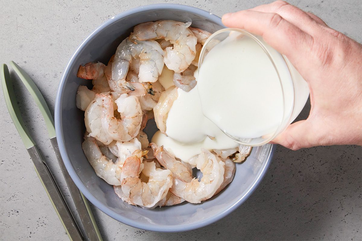 Overhead horizontal image of raw shrimp being coated with buttermilk in a mixing bowl.