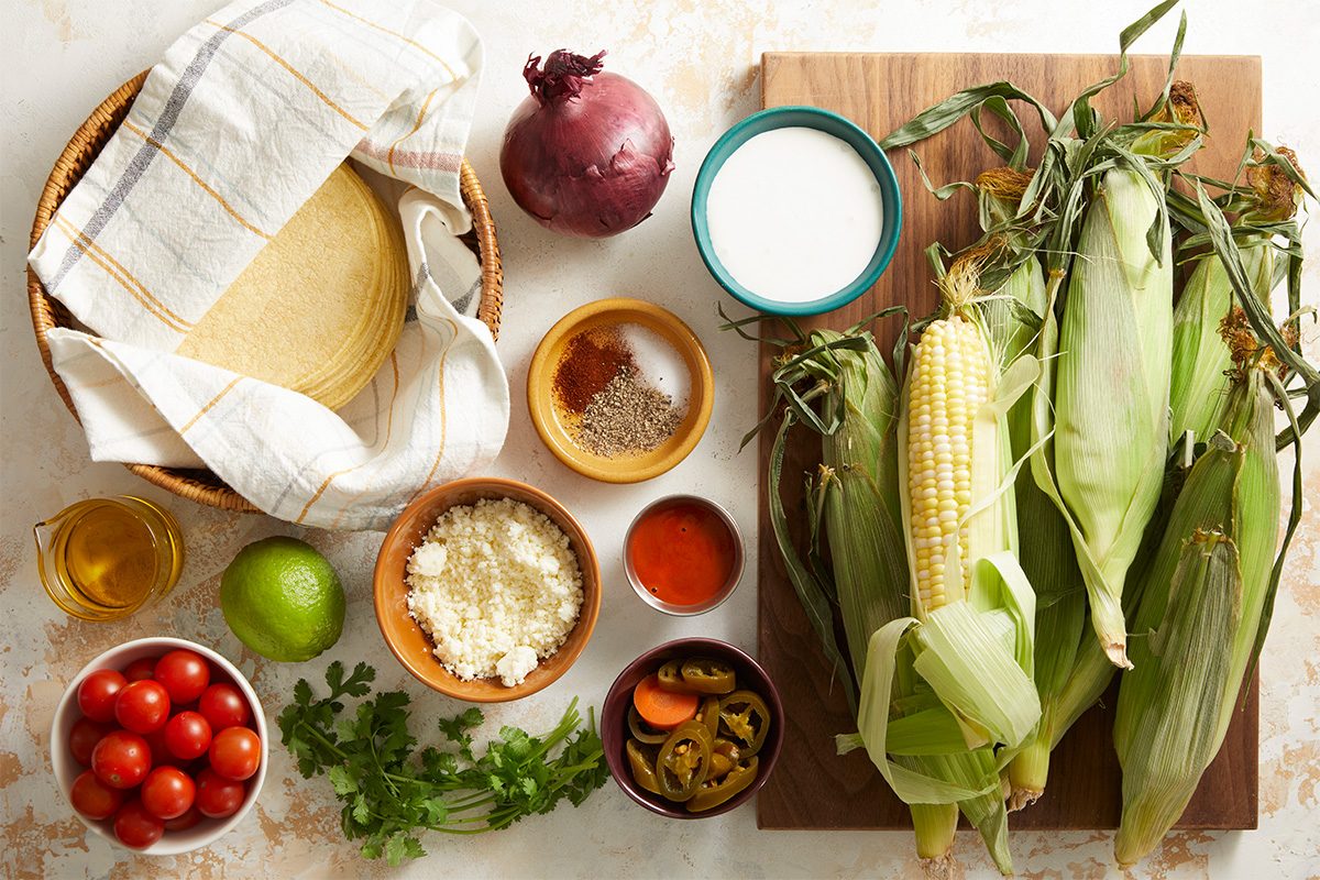 Overhead shot of fresh taco ingredients including corn on the cob, tomatoes, onion, cilantro, and cheese arranged neatly on a wooden surface with a tortilla and small bowls.