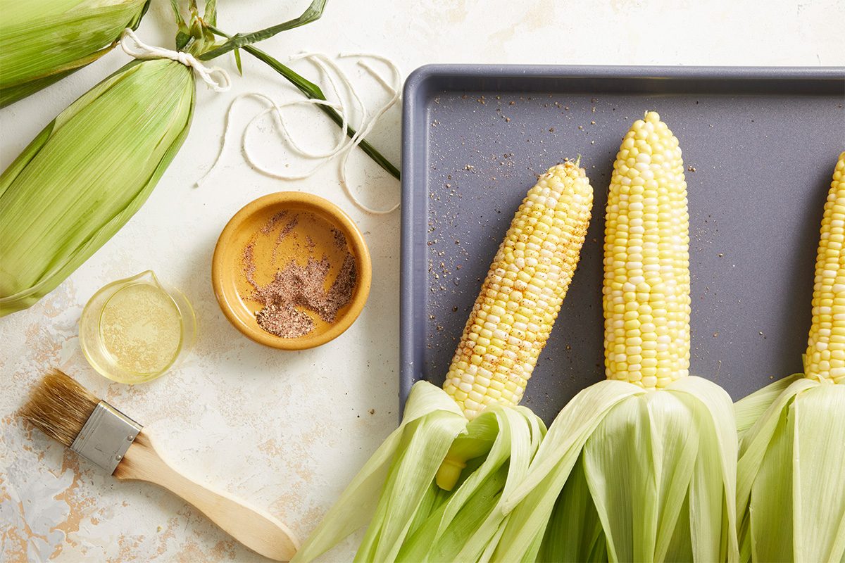 Overhead shot of partially husked corn cobs arranged on a metal baking sheet with a small bowl of seasoning and a brush on the side.