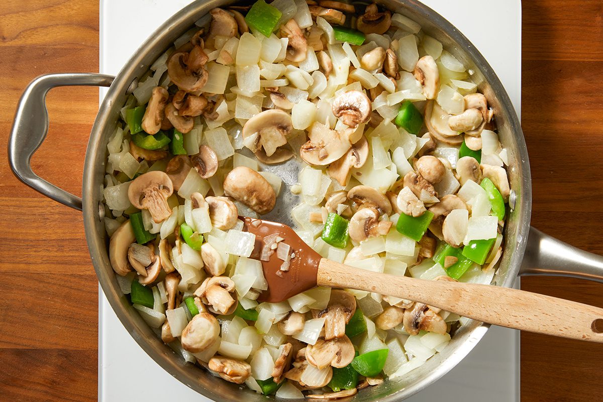 A pan on a stove filled with sautéed mushrooms, chopped onions, and green bell peppers being stirred with a wooden spoon.