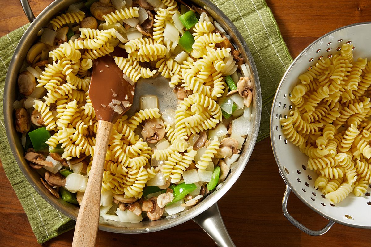 A pan with rotini pasta, chopped green bell peppers, onions, and mushrooms being stirred with a spatula sits next to a colander containing more cooked rotini, all on a green cloth.