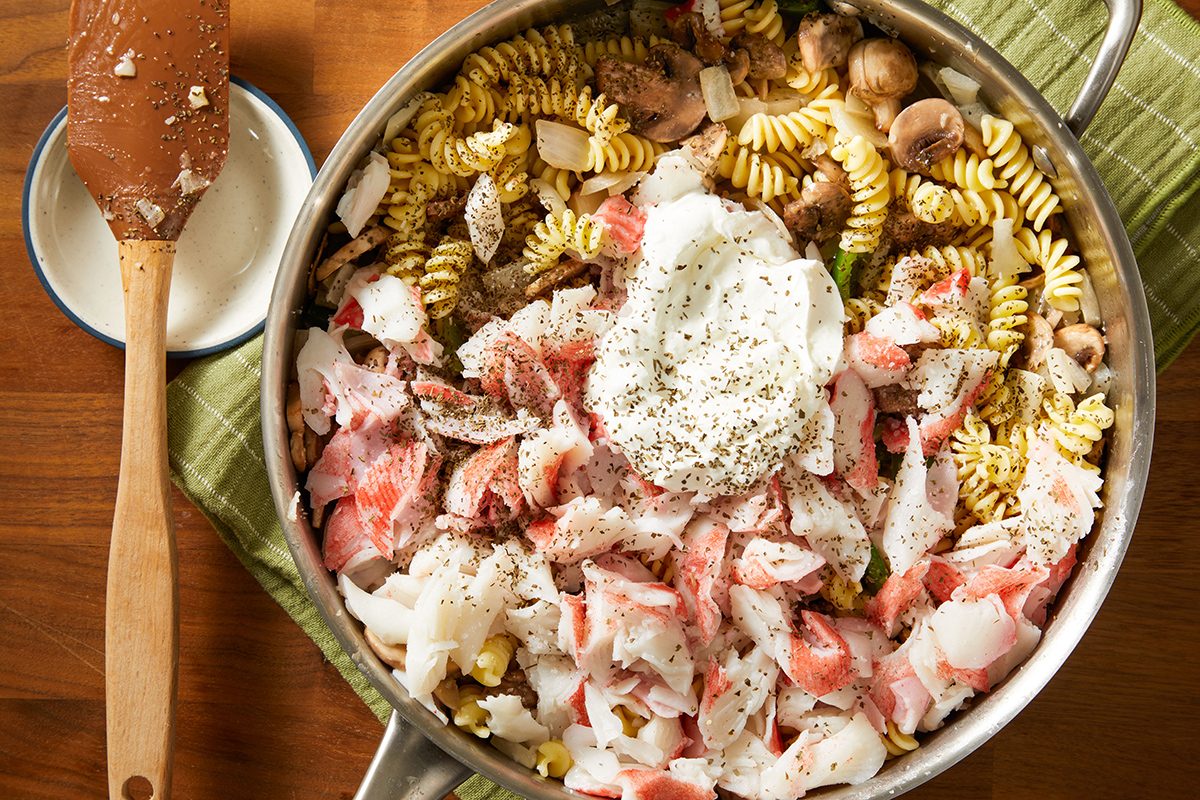A skillet filled with rotini pasta, chopped imitation crab, mushrooms, onions, and a dollop of creamy sauce, all seasoned with herbs, sits on a green cloth next to a wooden spoon.