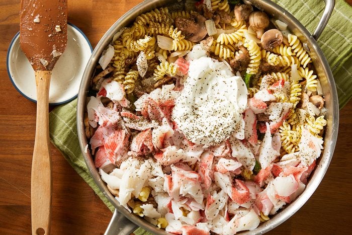 A skillet filled with rotini pasta, chopped imitation crab, mushrooms, onions, and a dollop of creamy sauce, all seasoned with herbs, sits on a green cloth next to a wooden spoon.
