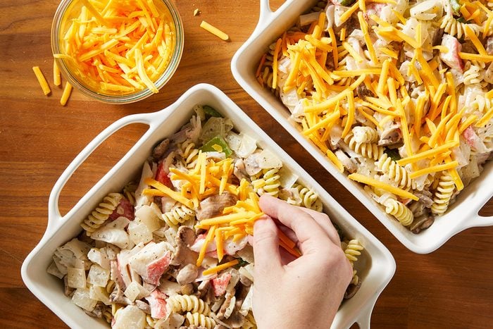A hand sprinkles shredded cheddar cheese over a casserole dish filled with rotini pasta, chopped meat, and vegetables. A small bowl of extra shredded cheese sits nearby on a wooden surface.