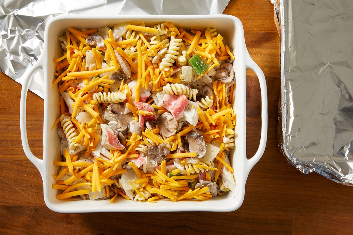 A white baking dish filled with a mixture of rotini pasta, chopped vegetables, and shredded cheese, sits on a wooden table next to a foil-covered dish.