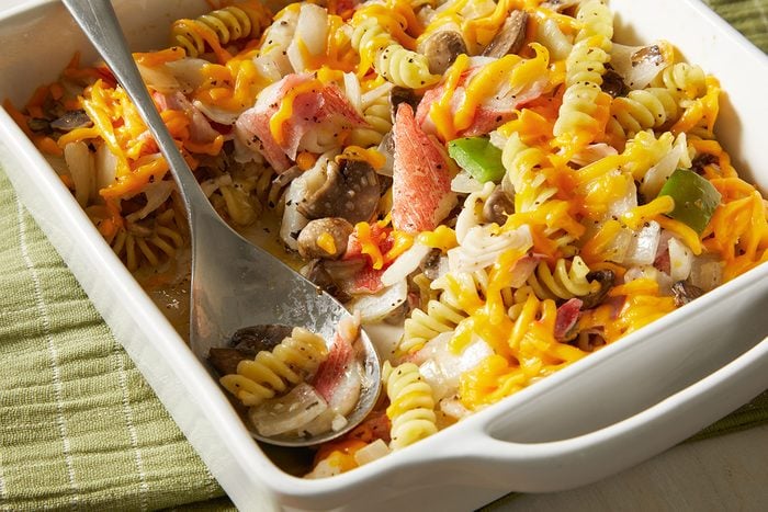 A white baking dish filled with a colorful pasta casserole featuring rotini noodles, shredded cheddar cheese, sliced mushrooms, green bell peppers, onions, and pieces of cooked meat. A large serving spoon rests inside.