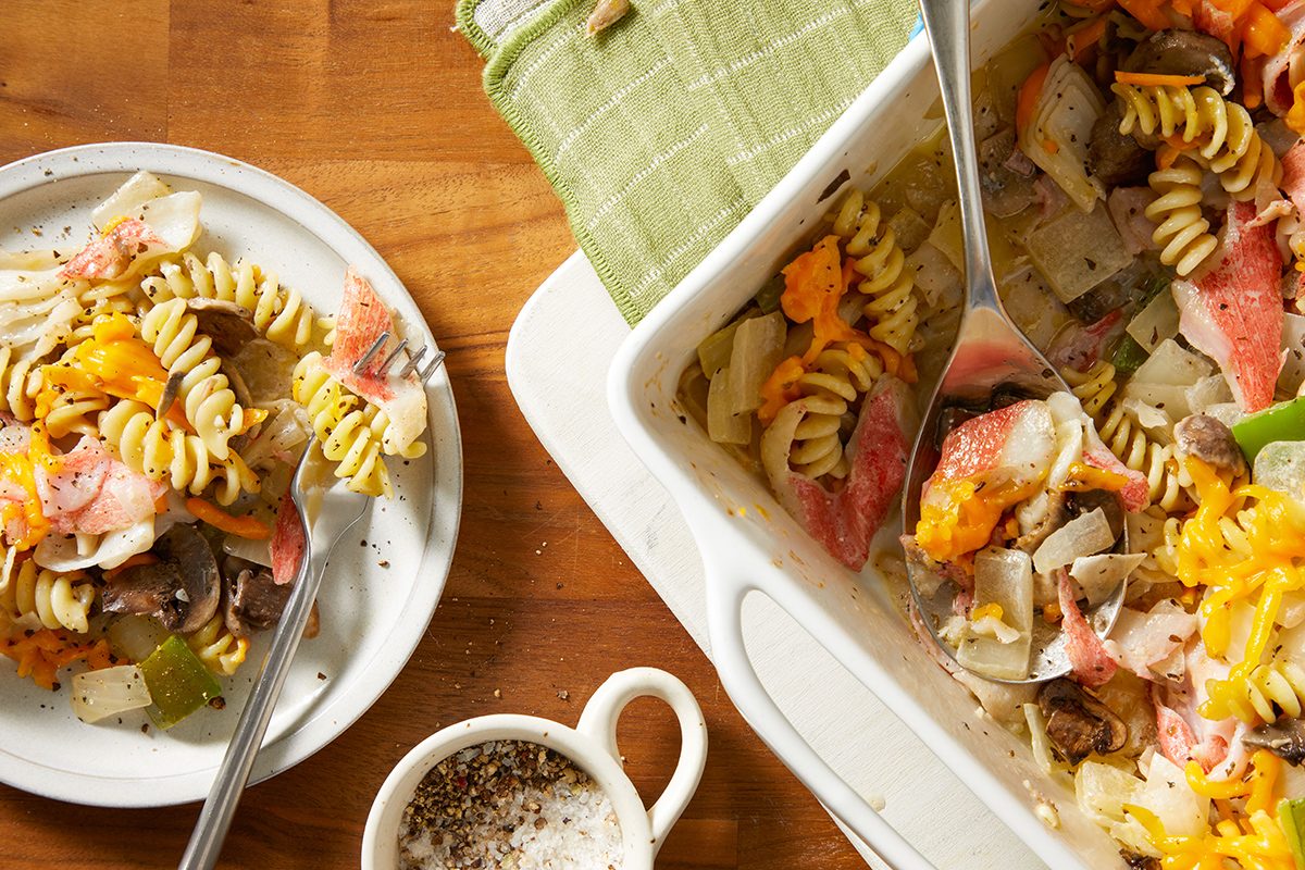 A serving of rotini pasta casserole with vegetables, cheese, and sliced meat on a white plate beside a baking dish, with a green cloth and a cup of black pepper on a wooden table.