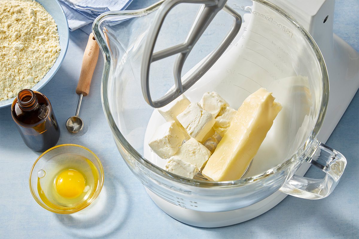 Overhead view of baking ingredients prepared in a stand mixer bowl, featuring softened butter and cream cheese ready for mixing, with egg and vanilla nearby. Clean, light blue background styled for a step-by-step baking tutorial for Death by Butter Cookies.
