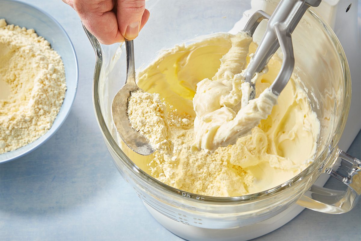 Overhead step-by-step baking image showing butter and cream cheese being mixed together in a stand mixer, capturing the creaming process for soft butter cookies. Styled on a light blue surface for a clean, modern recipe look.