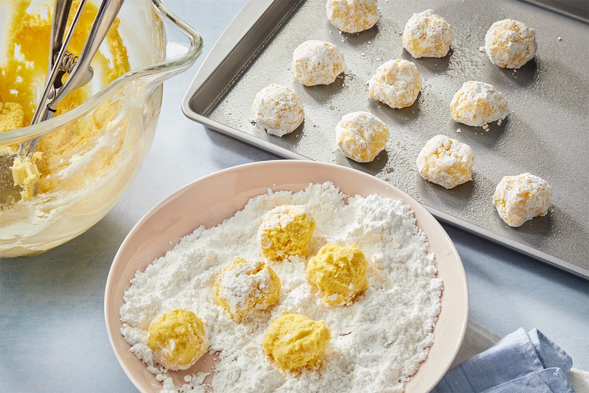 Overhead view of cookie dough balls rolled in confectioners’ sugar and arranged on baking trays, illustrating a key preparation step for Death by Butter Cookies before baking.